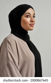 Happy young arabic woman in black hijab posing sideways and looking aside on light background, vertical shot. Beautiful muslim lady