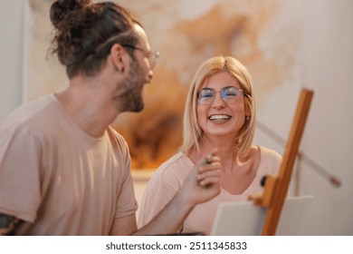 Happy young artistic couple standing at atelier with paintbrushes in hands and smiling at the camera. Portrait of a cheerful artist couple standing at creative art studio and bonding while painting. - Powered by Shutterstock