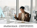 Happy young Asian business man celebrating success at work in office looking at laptop excited about financial trading growth, goals achievement good online results, screaming yes watching game.