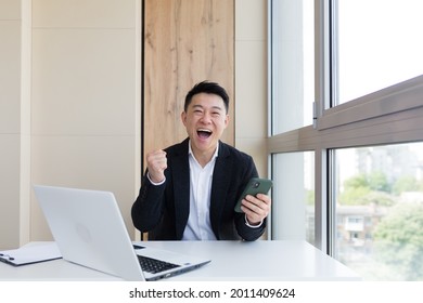 Happy Young Asian Business Man In Office, Looking At Camera, With Emotions Of Winner Or Victory, Financial Promotion Sports Betting. The Man Exclaims Happily While Playing The Game. Excited With Joy, 