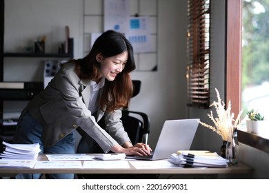 Happy Young Asian Businesswoman Standing Using Calculator And Laptop Computer At Office.
