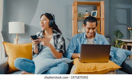 Happy Young Asian Couple Activity Man Use Laptop Computer Work Relax Enjoy With Women Wear Headphones Use Smartphone Listen Music At Sofa In Living Room At House. Young Married Work From Home Concept.