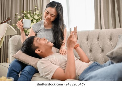 A happy young Asian couple enjoys talking and relaxing on a sofa in the living room together, the girlfriend is drinking coffee while her boyfriend lies on her lap. - Powered by Shutterstock