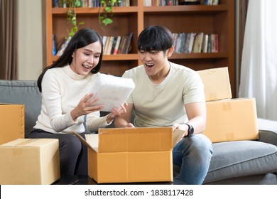 Happy Young Asian Couple Exciting With Goods Or Product After Opened Cardboard Box Or Parcel, Smiling Family Couple Sitting And Opening Carton Parcel In Living Room At Home. Delivery, Shipping Online 