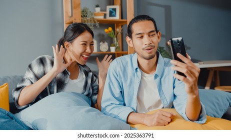 Happy Young Asian Couple Man And Woman Sit Couch Use Smartphone Facetime Video Call With Friends And Family In Living Room At Home. Stay At Home Quarantine, Social Distancing, Young Married Concept.