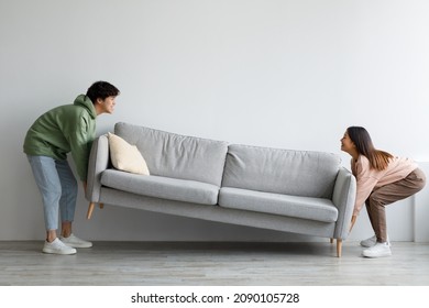 Happy Young Asian Couple Moving Sofa In Living Room, Replacing Furniture At Home, Copy Space. Millennial Husband And Wife Making Rearrangement In Their Apartment, Side View