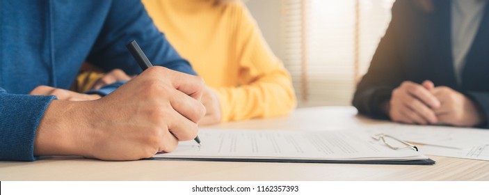 Happy Young Asian Couple And Realtor Agent. Cheerful Young Man Signing Some Documents While Sitting At Desk Together With His Wife. Signing Good Condition Contract. Panoramic Banner Background.