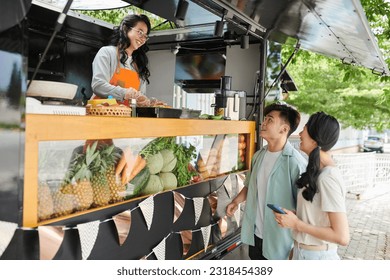 Happy young Asian couple standing by street food truck and looking at smiling female vendor in uniform preparing hotdogs for customers - Powered by Shutterstock