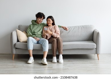 Happy Young Asian Couple Using Cellphone, Browsing Internet, Chatting Online, Relaxing On Sofa At Home, Free Space. Cheery Millennial Family Checking New Mobile Application, Resting On Couch Together