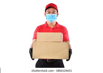 Happy Young Asian Delivery Man In Red Uniform, Medical Face Mask, Protective Gloves Carry Cardboard Box In Hands Isolated On White Background. Delivery Guy Give Parcel Shipment. Safe Delivery