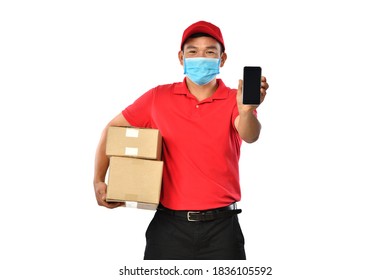 Happy Young Asian Delivery Man In Red Uniform, Medical Face Mask, Protective Gloves Carry Cardboard Box Showing Mobile Phone Isolated On White Background