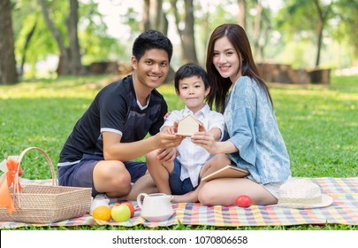 Happy Young Asian Family Having Fun Outside With Holding Home Together And Sitting Picnic In The Park. 