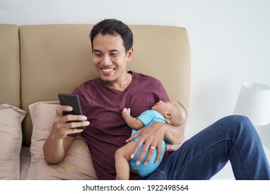 Happy Young Asian Father Holding His Newborn Sweet Adorable Baby Sleeping On His Arms While Using Mobile Phone On The Bed