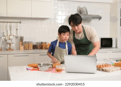 Happy young Asian father and kid are using a laptop computer to learn how to cooking food in their home kitchen. - Powered by Shutterstock