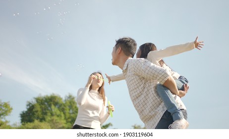 Happy Young Asian Father Piggyback With His Little Girl While Mother Run To Play With Her In The Park And Enjoyed Ourselves Together In The Green Garden. Family Enjoying Sunny Fall Day In Nature.