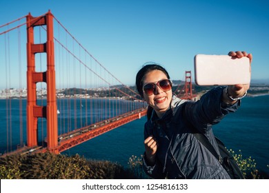 Happy young asian female tourist taking selfie in San Francisco by Golden Gate Bridge USA. Interracial modern woman using smart phone taking self portrait wearing sunglasses with blue sky and sea. - Powered by Shutterstock