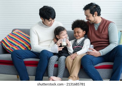 Happy Young Asian Gay Couple With Diverse Adopted Children, African And Caucasian, Drinking Milk And Sitting On Sofa At Home. Older Boy With Milk Mustache Cheer Up Little Girl Drink Milk. Lgbt Family