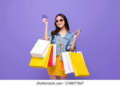 Happy Young Asian Girl Carrying Colorful Shopping Bags With Credit Card And Mobile Phone In Hands Isolated In Purple Background