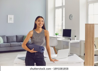 Happy Young Asian Girl Holding Yoga Mat And Looking At Camera Leaning On Office Desk. Smiling Woman Looking Forward To Fitness Workout And Getting Ready To Go To The Gym After A Hard Day At Work