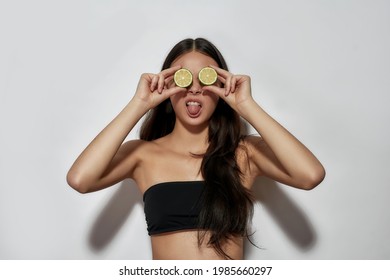 Happy Young Asian Girl Holding Lime Slices Near Her Eyes While Posing On Light Background And Fooling At Camera. Skincare Concept, Beauty And Wellness