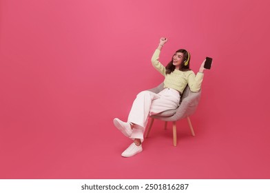 Happy young asian girl listening music on mobile phone with headphone dancing on chair isolated on pink background. People lifestyle concept - Powered by Shutterstock