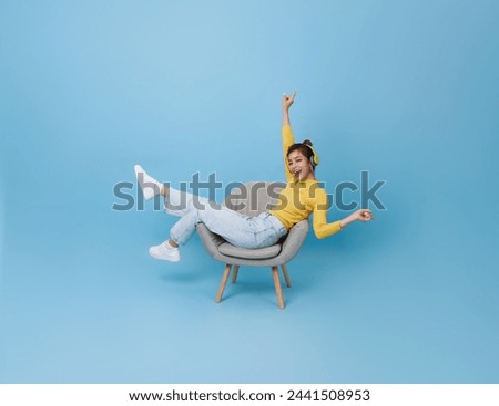 Happy young asian girl listening music with headphone dancing on chair isolated on blue background. People lifestyle concept