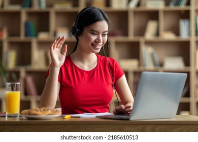 Happy Young Asian Girl Student In Headphones Studying With Laptop, Greet, Waving Hand, Watching Online Lesson In Room Interior. Client Support, Education At Home, Work And Knowledge During Covid-19