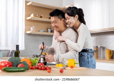 Happy Young Asian Lady Hugging Her Spouse Cooking Dinner, Cheerful Chinese Husband And Wife Preparing Healthy Meal Together At Home, Cozy Kitchen Interior, Side View, Copy Space