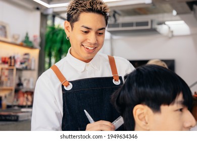 Happy young Asian male hairdresser smiling wearing apron in modern salon holding brush and scissors trimming hair of client - Powered by Shutterstock