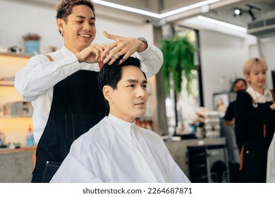 Happy young Asian male hairdresser smiling wearing apron in modern salon holding brush and scissors trimming hair of client - Powered by Shutterstock