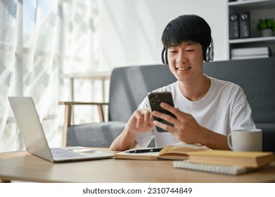 A happy young Asian man is listening to music through his headphones and scrolling on social media through his phone at a coffee table in the living room. - Shutterstock ID 2310744849