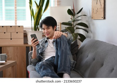 Happy young asian man lying on couch at home and using smart phone.