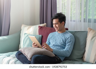 Happy Young Asian Man Sitting On The Sofa Watching A Tablet At Home.