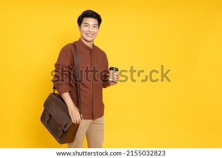 Happy young Asian man standing with leather bag and hot coffee cup isolated on yellow background