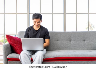 Happy Young Asian Man Using Laptop Computer Sitting O Sofa In Living Room, Work From Home Concept.