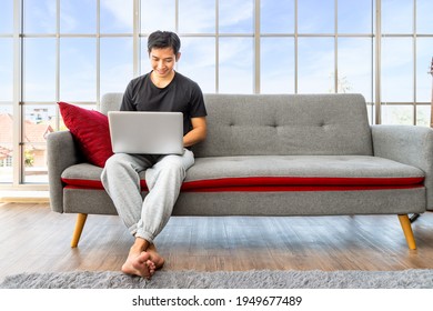 Happy young Asian man using laptop computer sitting o sofa in living room, work from home concept.