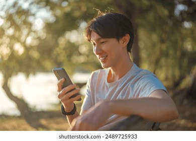 Happy young asian man using smartphone while sitting on bench in public park.