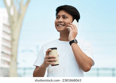 Happy young asian man wearing white t-shirt talking on smartphone, chatting with friend, set up date, drinking takeaway coffee, posing at urban city area, looking at copy space and smiling - Powered by Shutterstock