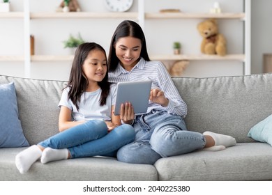 Happy young asian mom and daughter using digital tablet, watching videos or surfing internet, sitting on sofa at home, copy space. Young korean family using pad together