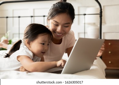 Happy Young Asian Mom And Little Ethnic Daughter Lying In Bed At Home Watch Video On Modern Laptop, Smiling Vietnamese Mother Or Nanny Relax In Bedroom With Small Biracial Girl Child Using Computer