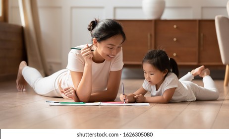 Happy Young Asian Mother And Cute Little Biracial Daughter Lying On Warm Wooden Floor In Living Room Painting Together, Millennial Ethnic Mom Or Nanny Relax With Small Vietnamese Girl Child Drawing