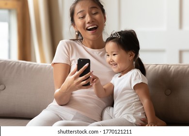 Happy Young Asian Mother Showing Funny Educational Application On Mobile Phone To Interested Cute Small Kid Daughter, Relaxing Together On Sofa, Excited Family Using Digital Gadget, Watching Videos.