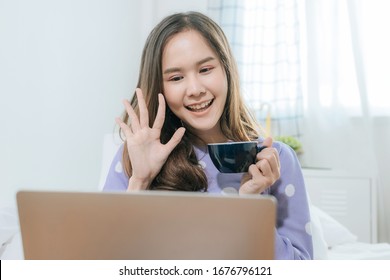 happy young asian pretty woman drinking coffee and video call chat on digital laptop computer and sitting on bed at home at morning, facetime connection, internet, quarantine, technology concept