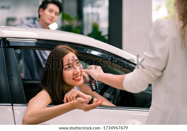 Happy Young Asian smiling woman getting keys of a\
new car. Beautiful Asian Woman is choosing a new car at dealership.\
Salesman giving a new car keys to beautiful owners. car sale,car\
rental concept