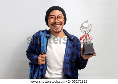 Happy young Asian student, adorned in a beanie hat, eyeglasses, and a casual shirt, proudly lifts his trophies while smiling and looking directly at the camera, while standing against white background