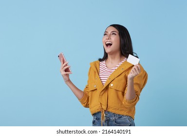 Happy Young Asian Teen Woman Laughing Holding Mobile Phone And Credit Card Isolated On Studio Blue Background.