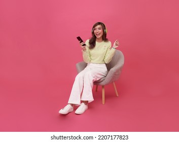 Happy young Asian teen woman smiling listening music in headphones and sitting on chair isolate on pink background. - Powered by Shutterstock