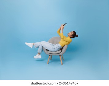 Happy young Asian teen woman using mobile phone. While her sitting relax on chair isolated on blue background. - Powered by Shutterstock