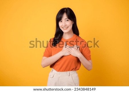 Happy young Asian woman 30s, wearing an orange shirt, holds hands on chest on yellow background. healthcare wellness concept.