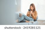 Happy young asian woman browsing surfing wireless internet on mobile smartphone while sitting a couch in living room at home, Shopping online via website